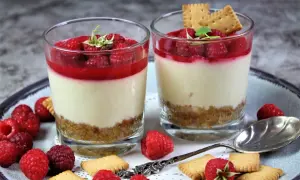 Zwei Gläser mit Himbeer-Dessert, Schicht aus Keksboden, Creme und Himbeersauce, garniert mit frischen Himbeeren und Keksen.