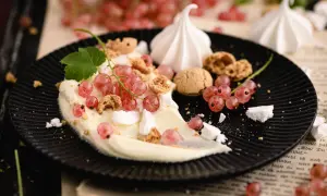 Cremiges Dessert mit roten Johannisbeeren, Baiser und Kekskrümeln auf schwarzem Teller.