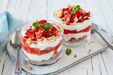 Zwei Gläser mit geschichtetem Erdbeer-Mascarpone-Dessert, garniert mit Minze und Keksbröseln.