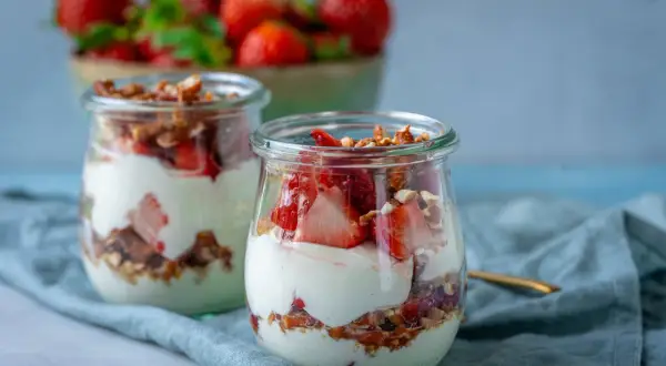 Erdbeer-Dessert im Glas mit Joghurt und Granola, im Hintergrund eine Schüssel mit Erdbeeren.