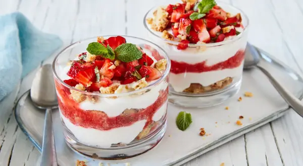 Zwei Gläser mit geschichtetem Erdbeer-Mascarpone-Dessert, garniert mit Minze und Keksbröseln.
