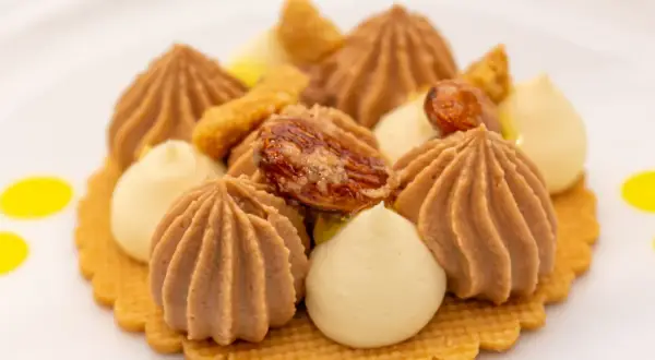 Ein kunstvolles Dessert mit braunen und hellen Cremetupfern, Nüssen und gelben Tupfern auf einem Keks.
