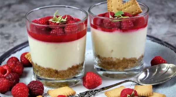 Zwei Gläser mit Himbeer-Dessert, Schicht aus Keksboden, Creme und Himbeersauce, garniert mit frischen Himbeeren und Keksen.