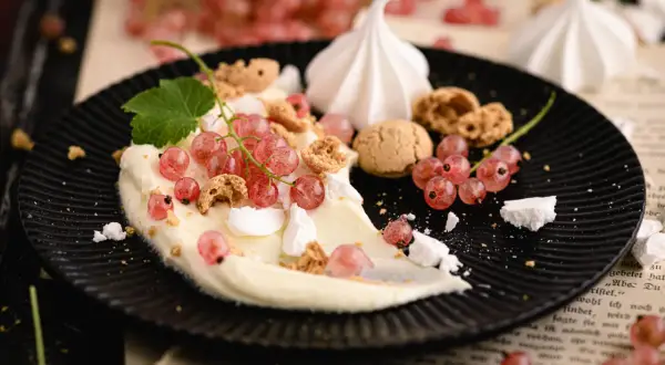 Cremiges Dessert mit roten Johannisbeeren, Baiser und Kekskrümeln auf schwarzem Teller.