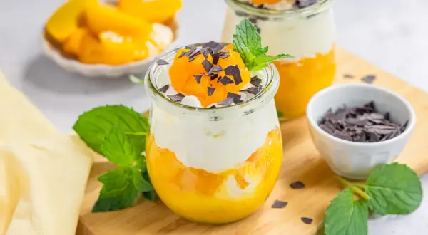 Zwei Gläser mit Schichtdessert aus Pfirsich und Sahne, garniert mit Schokostreuseln und Minze.