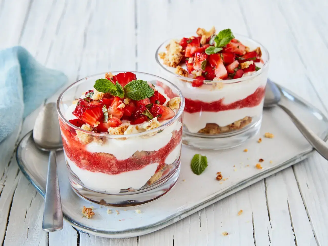 Zwei Gläser mit geschichtetem Erdbeer-Mascarpone-Dessert, garniert mit Minze und Keksbröseln.
