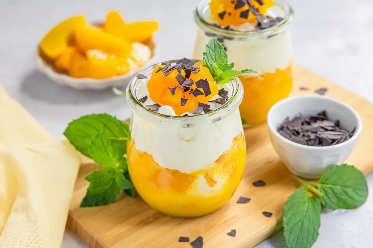 Zwei Gläser mit Schichtdessert aus Pfirsich und Sahne, garniert mit Schokostreuseln und Minze.