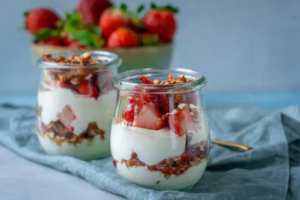 Erdbeer-Dessert im Glas mit Joghurt und Granola, im Hintergrund eine Schüssel mit Erdbeeren.
