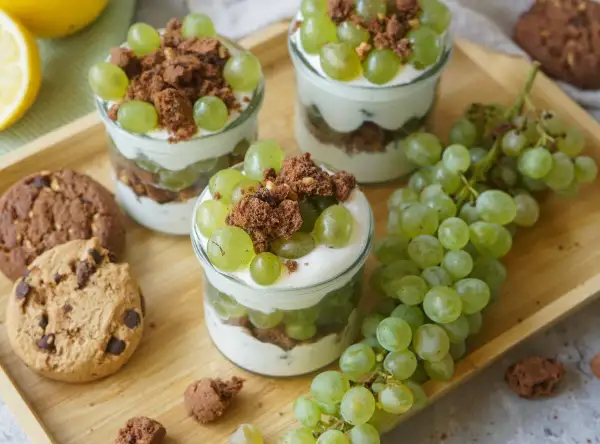 Drei Gläser mit Trauben-Keks-Dessert, garniert mit grünen Trauben und Schokostückchen, daneben Kekse und Trauben.