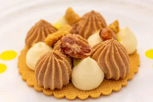 Ein kunstvolles Dessert mit braunen und hellen Cremetupfern, Nüssen und gelben Tupfern auf einem Keks.