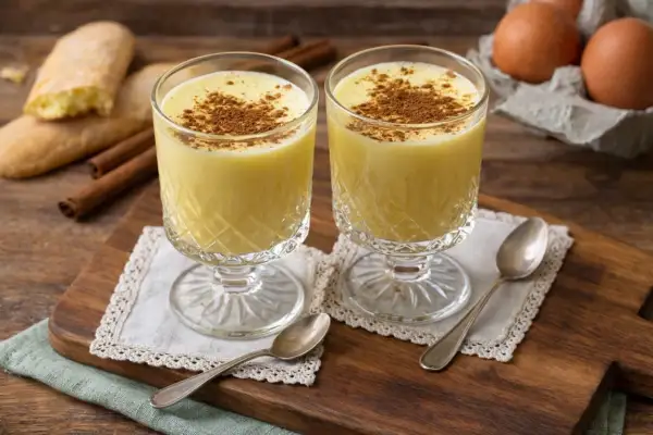 Zwei Gläser Zabaione mit Zimt bestreut, daneben Löffel, Kekse und Eier.