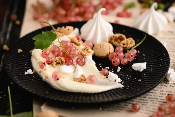Cremiges Dessert mit roten Johannisbeeren, Baiser und Kekskrümeln auf schwarzem Teller.